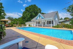 A swimming pool with a covered patio area, a white building with a blue roof, and a large tree in the background.