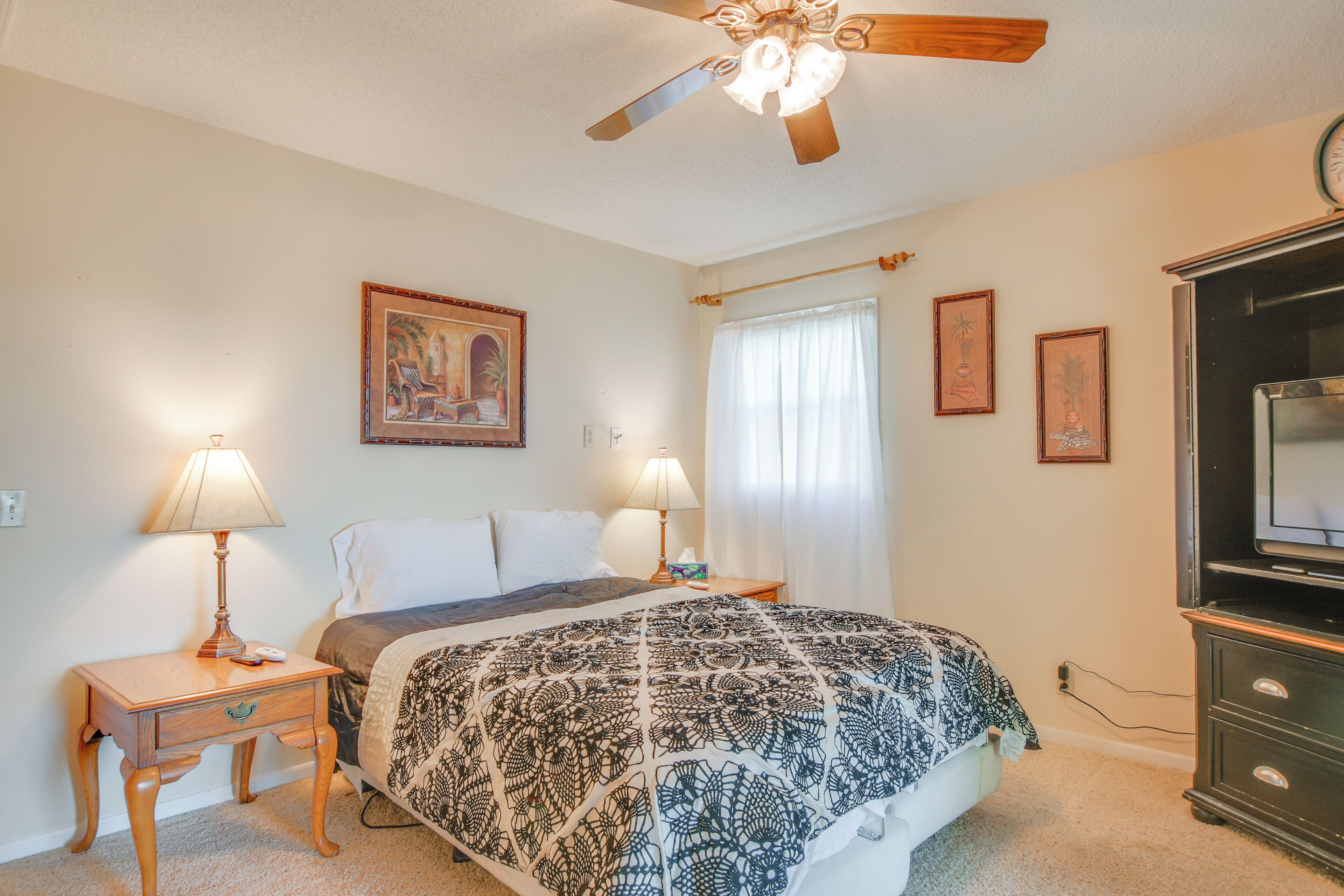 A bedroom with a bed, two bedside tables with lamps, a ceiling fan, a television, and framed pictures on the wall.
