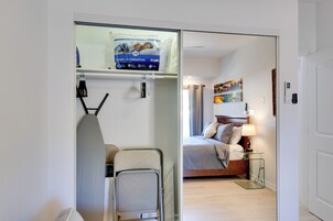 A hotel room with a bed, a bedside table with a lamp, a painting on the wall, and a closet with hanging clothes.