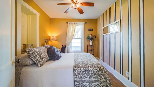 A bedroom with a bed, a ceiling fan, a window with curtains, a bedside table with a lamp, and a wall with vertical stripes.