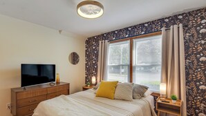 A bedroom with a bed, a TV on a wooden dresser, a nightstand with a lamp, and a window with curtains. - 1 Mi to Dtwn Burlington: Modern Home w/ Yard! (Burlington)