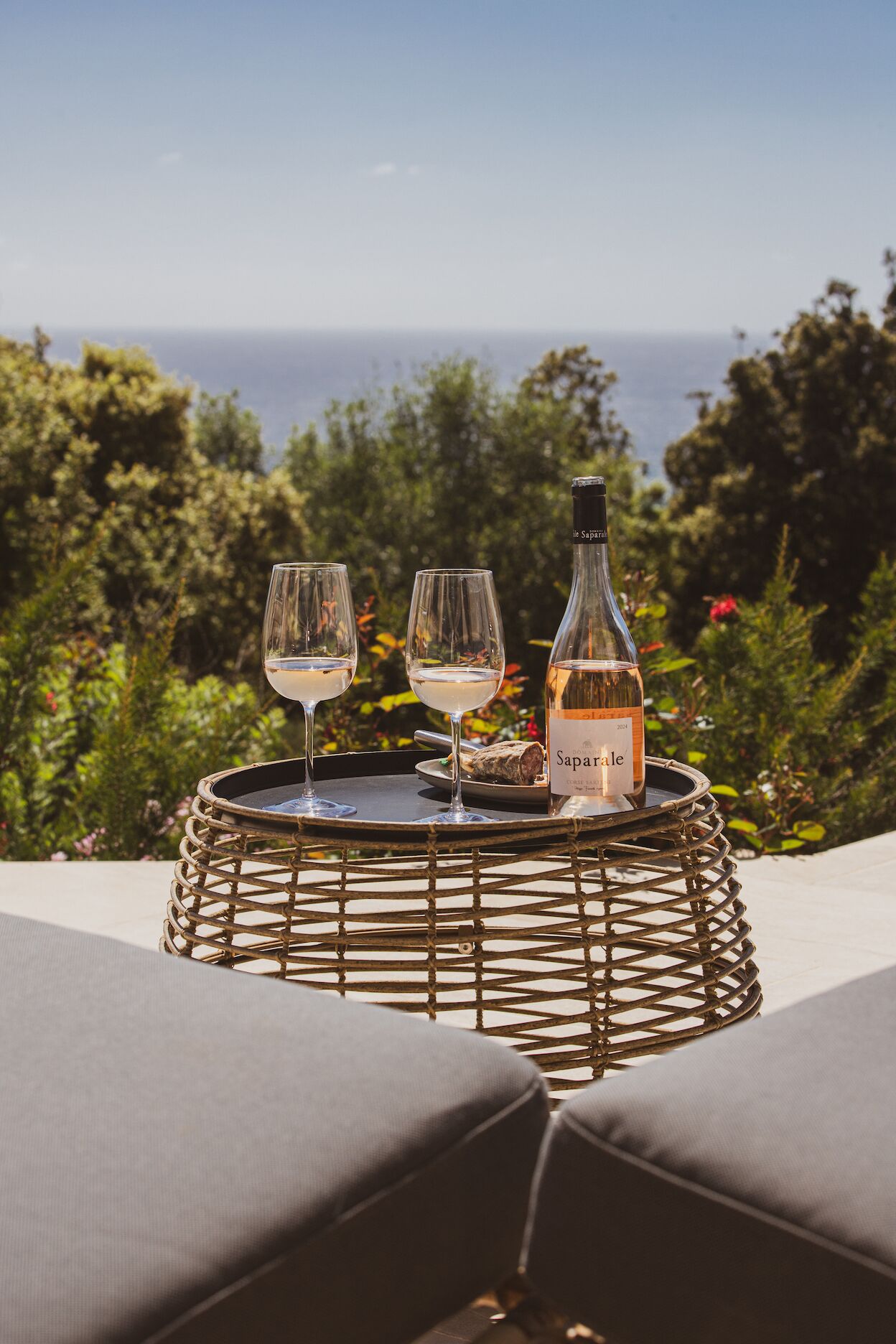 Een rieten tafeltje met een fles Sapparose wijn en twee glazen, op een terras met uitzicht op de oceaan.