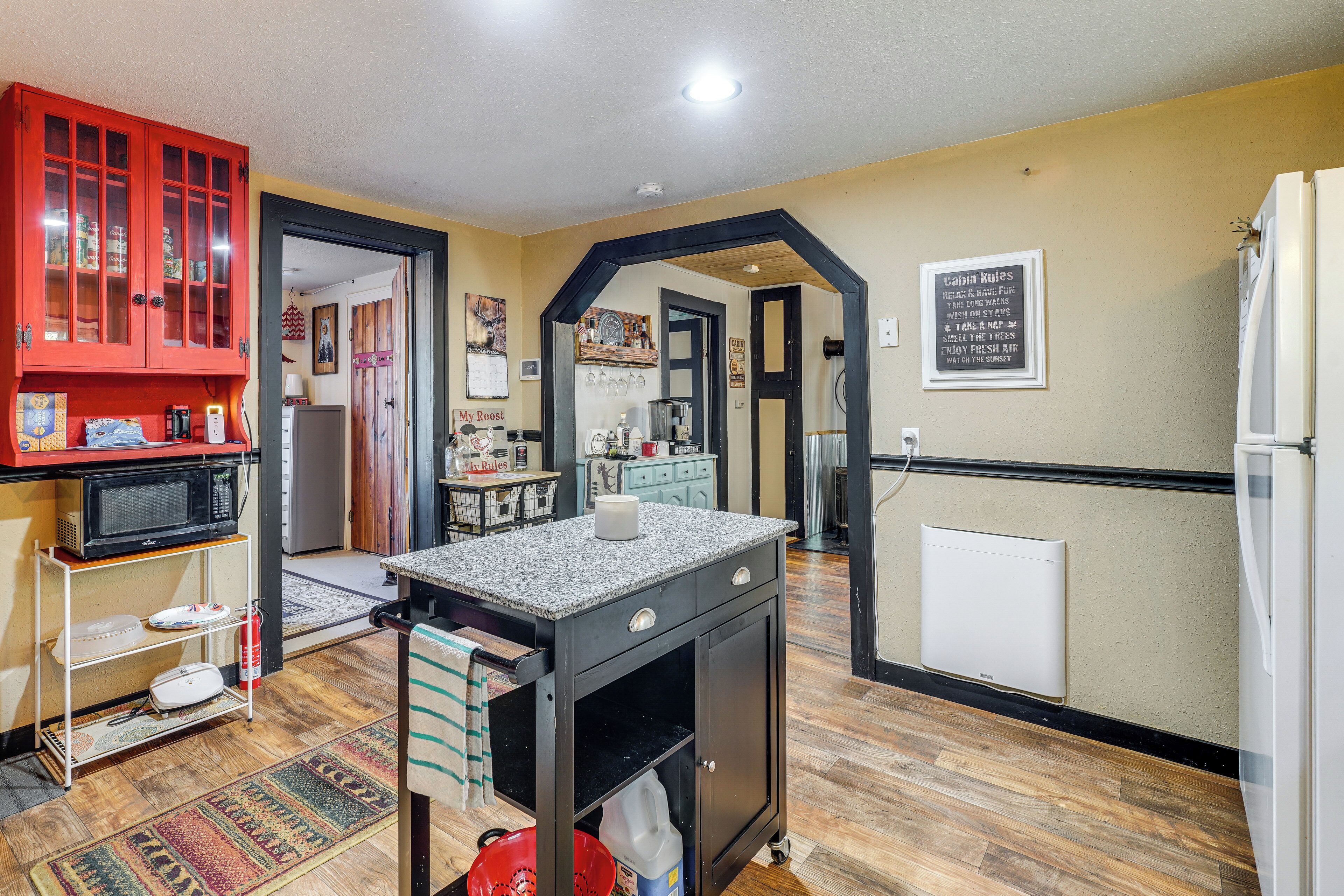 A kitchen with a granite countertop, a microwave, a refrigerator, and a red cabinet. There is a rug on the floor and a sign on the wall.