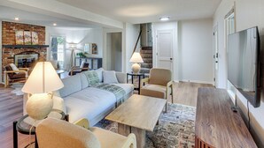 A spacious living room with a sofa, armchairs, a coffee table, and a TV. There is a fireplace with a brick surround and a lamp on a side table. - 19 Mi to Kansas City: Group Retreat w/ Game Room! (Blue Springs)