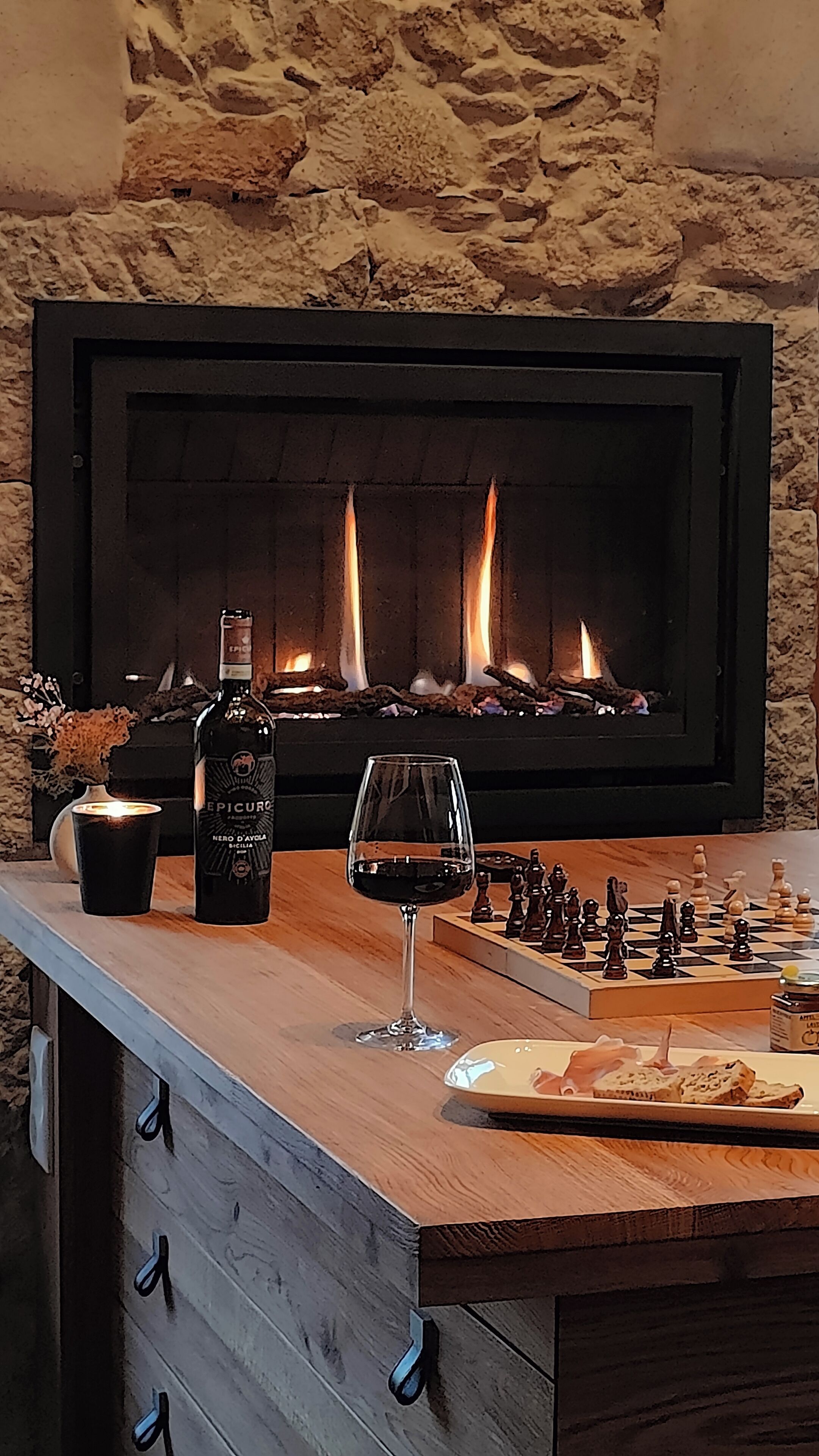 A fireplace with a stone wall backdrop, a bottle of wine, a glass of red wine, a chessboard, and a wooden bar counter.