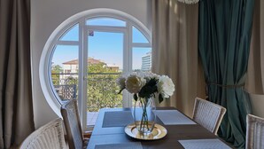 A dining area with a round table, chairs, a vase with flowers, a chandelier, and a view through a large window. - Apartamenty Świnoujście - Trzy Korony (Świnoujście)