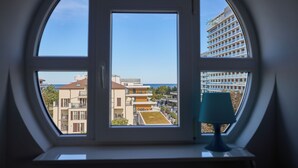A window with a view of a building complex, including a high-rise and lower buildings, through a rounded window frame. - Apartamenty Świnoujście - Trzy Korony (Świnoujście)