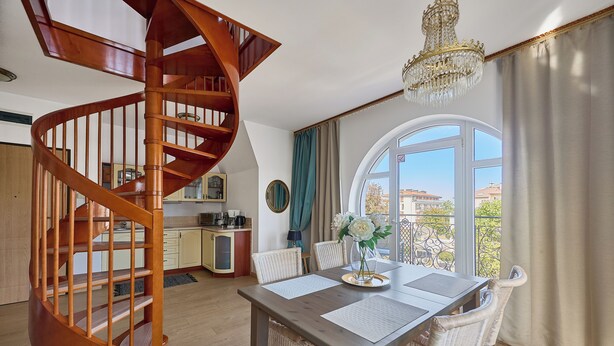 A modern interior with a wooden spiral staircase, a dining area with wicker chairs, and a large window offering a view of the city. - Apartamenty Świnoujście - Trzy Korony (Świnoujście)