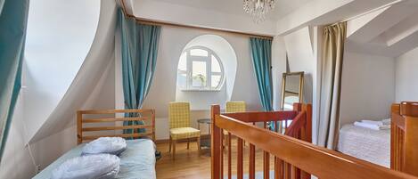 Un dormitorio con cama de madera, una mesita pequeña y una silla. Se ve una ventana con cortinas, un candelabro y un espejo en la pared.