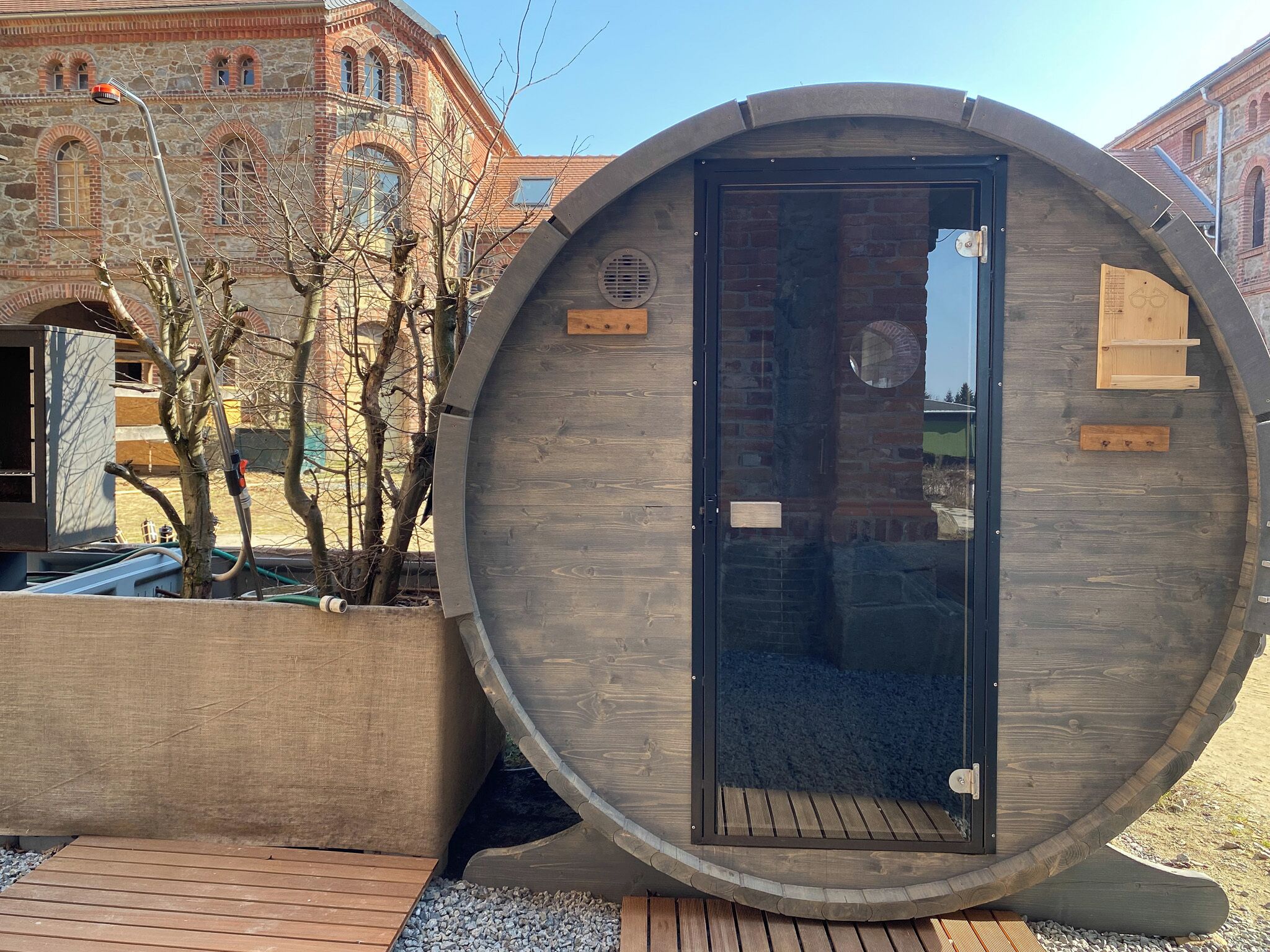 A wooden cylindrical structure with a door, positioned outdoors with a building in the background.