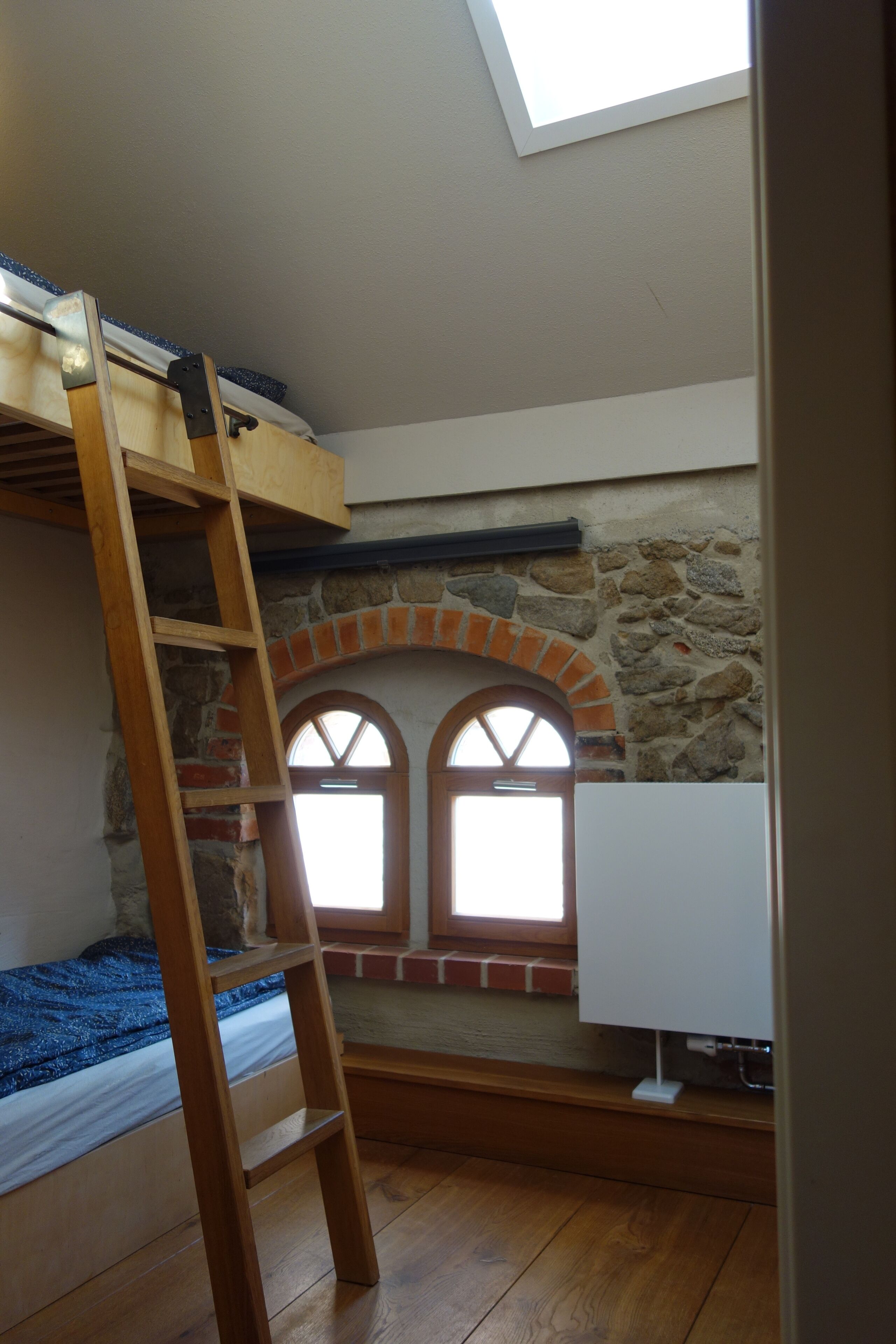 Ein Schlafzimmer im Stil eines Lofts mit einer Holzlade, einem Dachfenster und einer Steinwand mit Rundbogenfenstern.