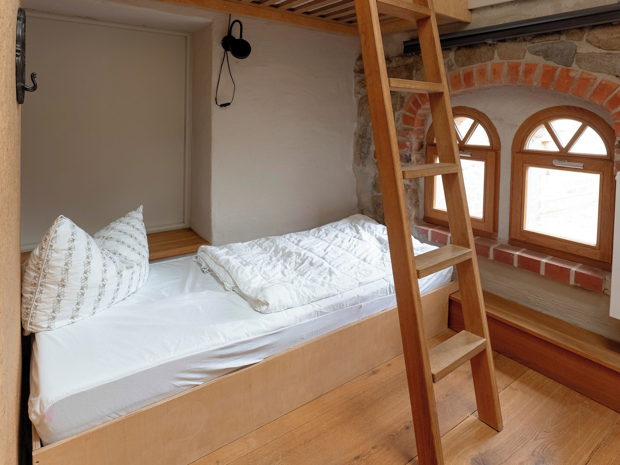 Un lit superposé avec un escalier en bois, une literie blanche et un mur de briques agrémenté de fenêtres en arc de cercle.