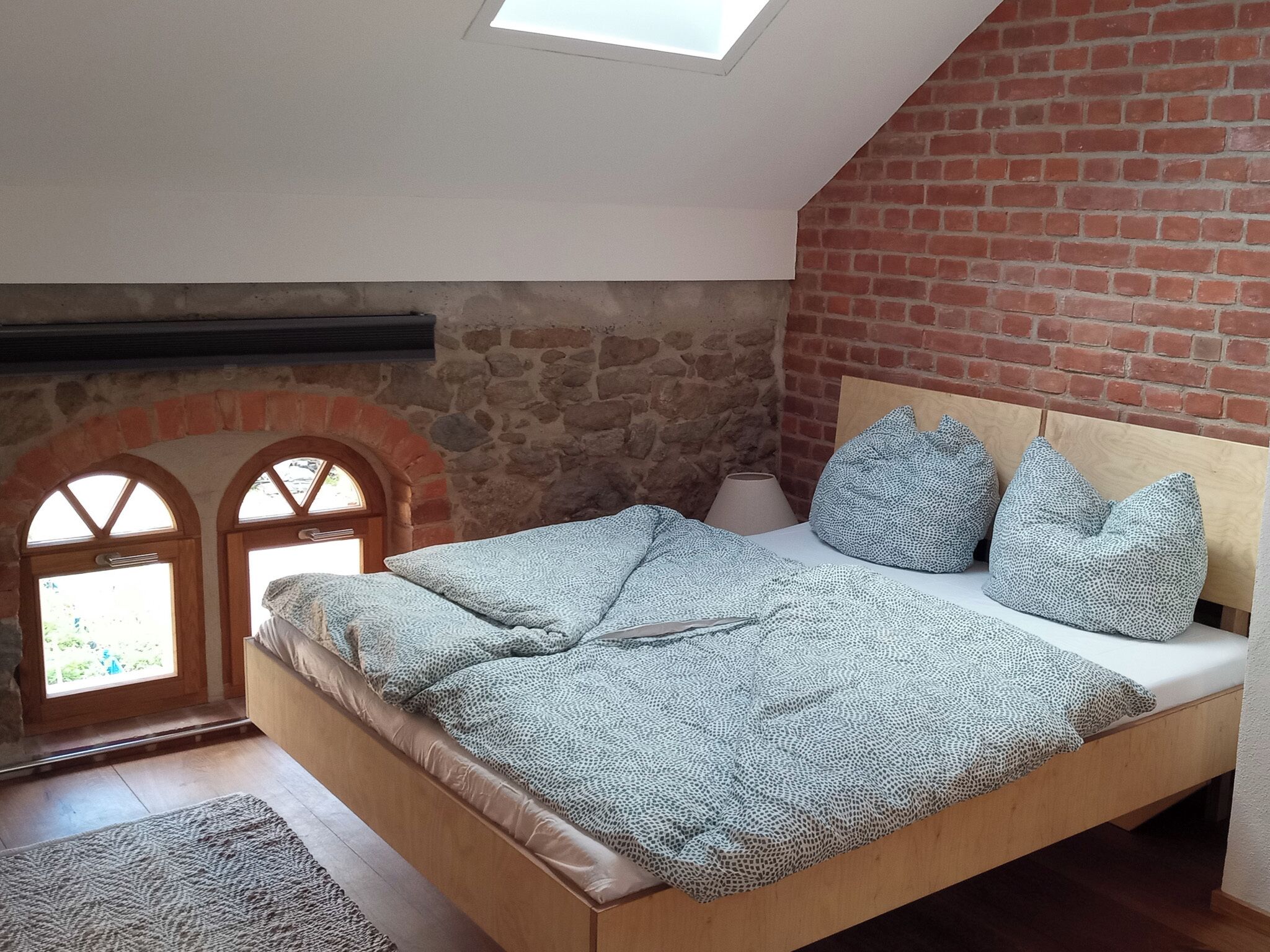 Un dormitorio con techo inclinado, pared de ladrillo, estructura de cama de madera y vista por una ventana con arco de piedra.