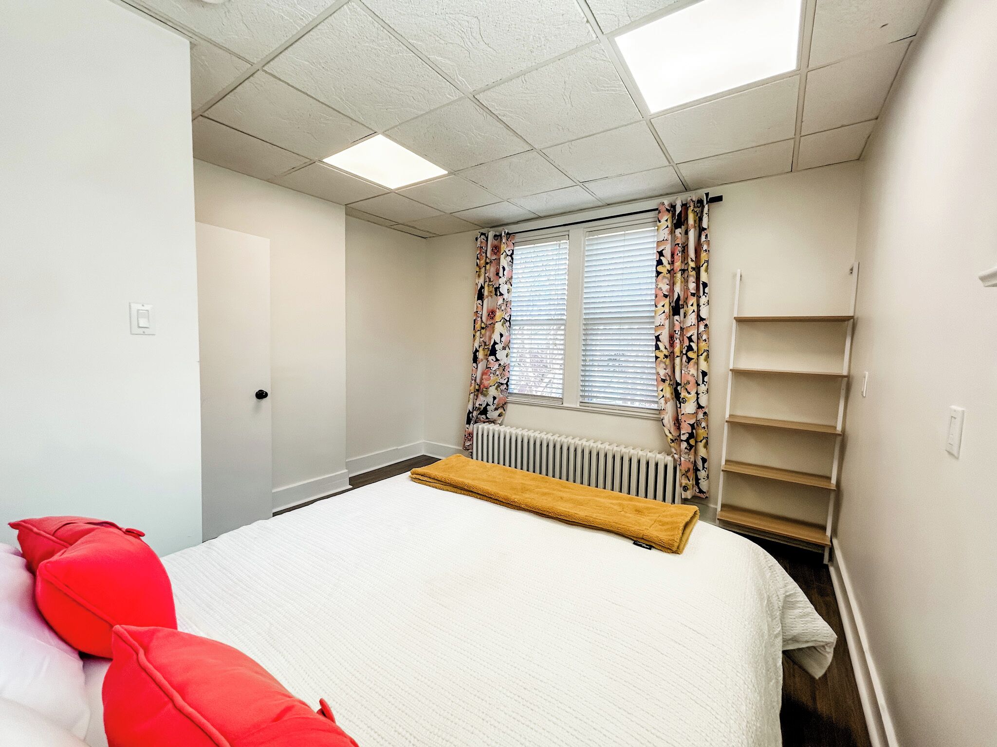 A bedroom with a bed, a radiator, a window with floral curtains, and a built-in shelf unit.