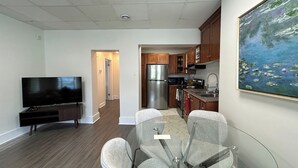 A modern living room with a flat-screen TV, a glass dining table with chairs, a kitchen area with wooden cabinets, and a painting on the wall.