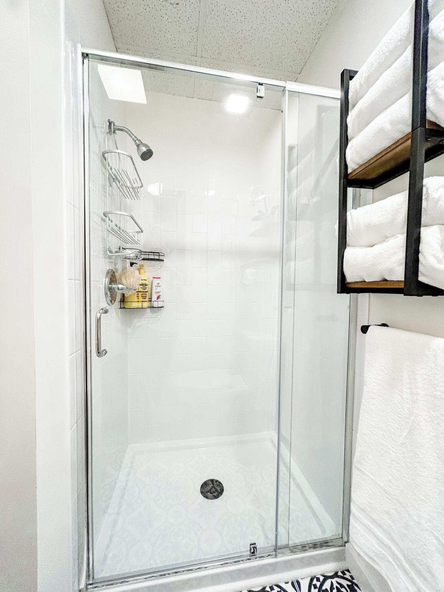 Ducha con cerramiento de vidrio, pared con azulejos blancos, puerta de vidrio y un perchero con toallas dobladas.