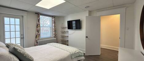 A bedroom with a bed, a TV mounted on the wall, a radiator, a window with curtains, and a door leading to another room.