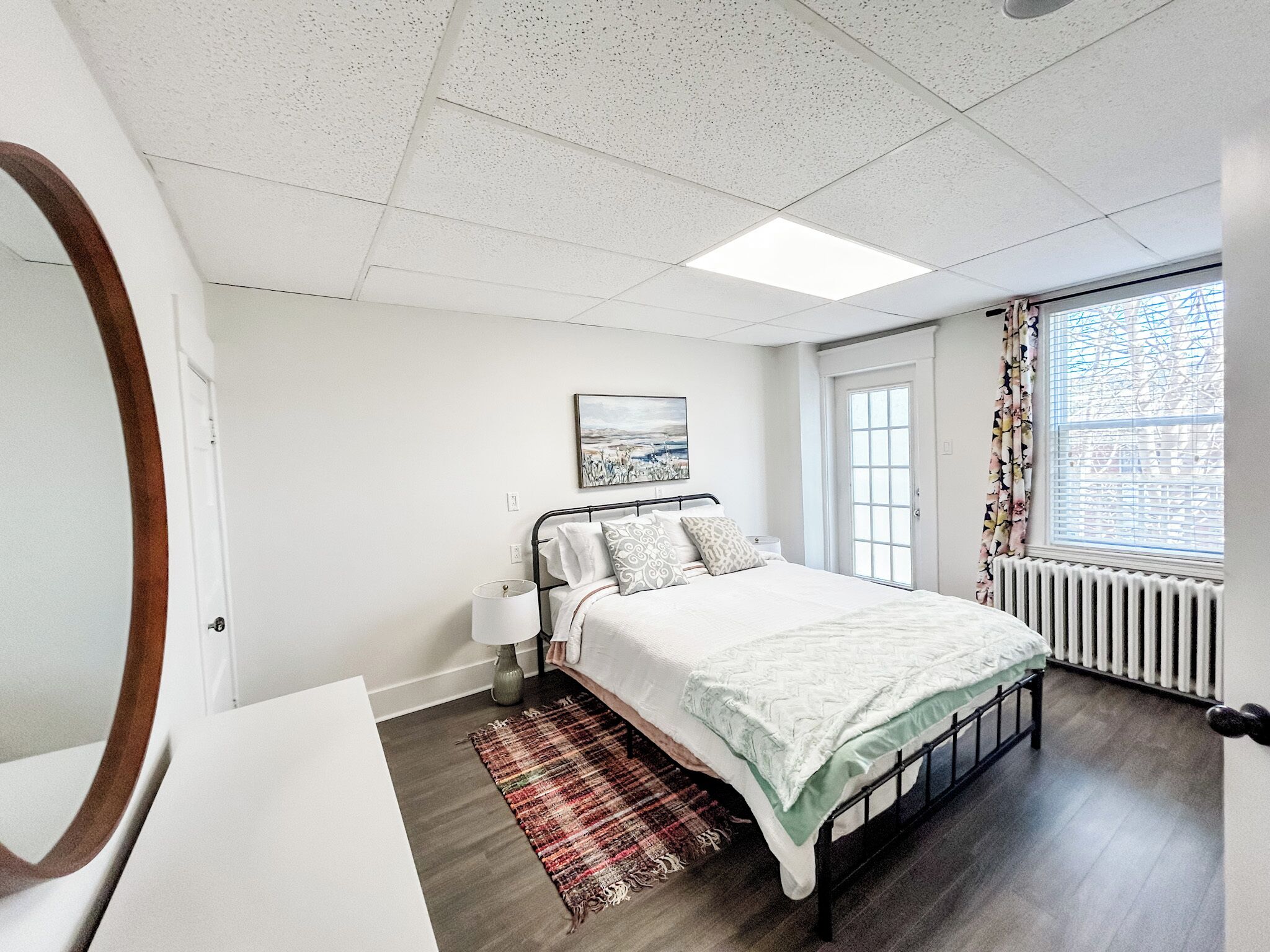 A bedroom with a bed, a bedside table, a lamp, a window with curtains, a radiator, and a wall-mounted picture.