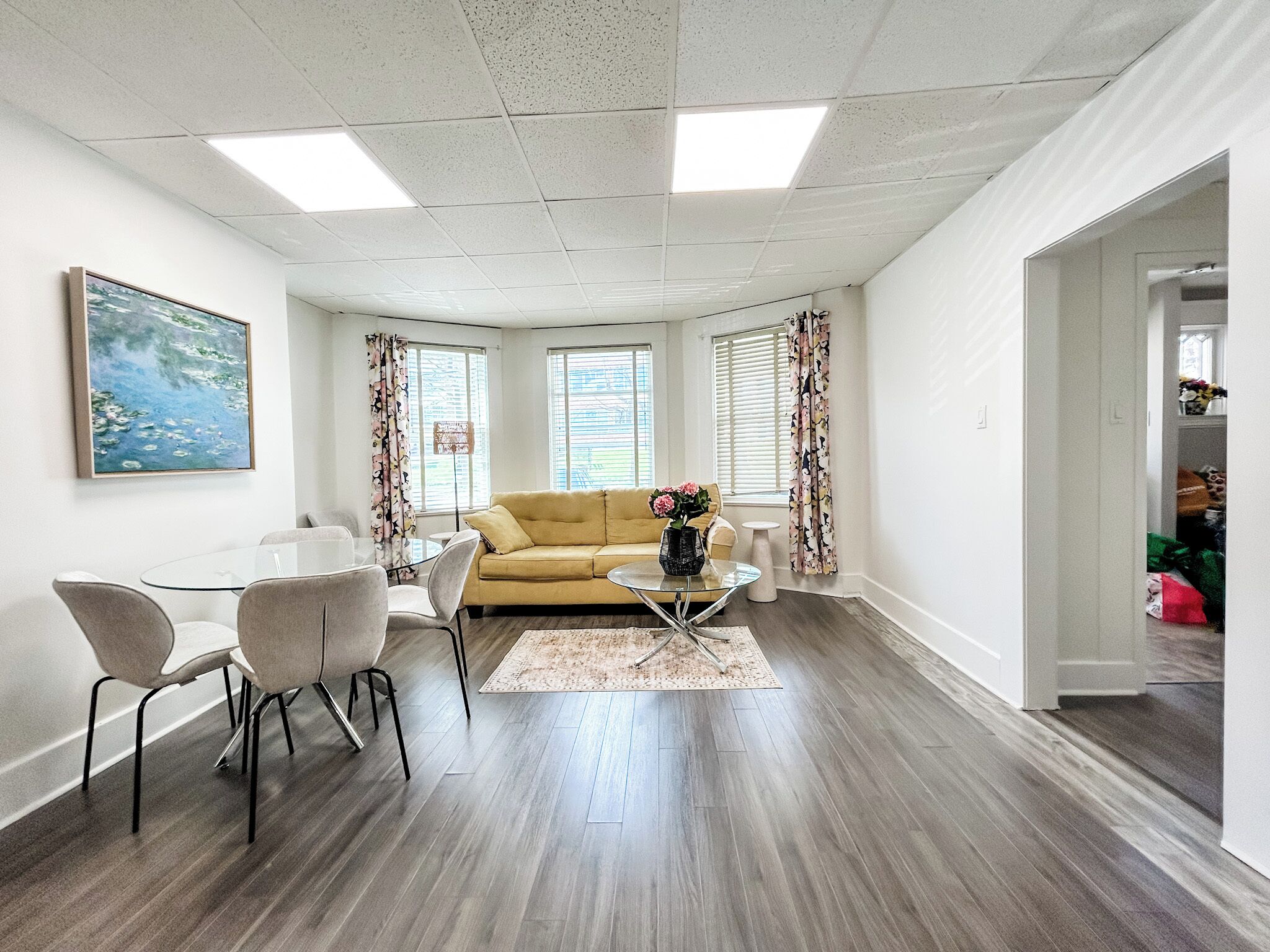Una sala moderna con un sofá, una mesita de centro y sillas. Hay ventanas con cortinas, un cuadro en la pared y una puerta que conduce a otra habitación.