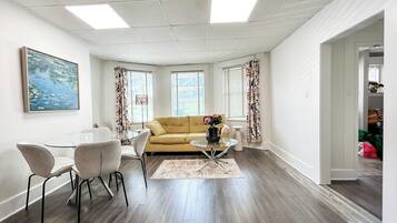 A modern living room with a sofa, coffee table, and chairs. There are windows with curtains, a painting on the wall, and a door leading to another room.