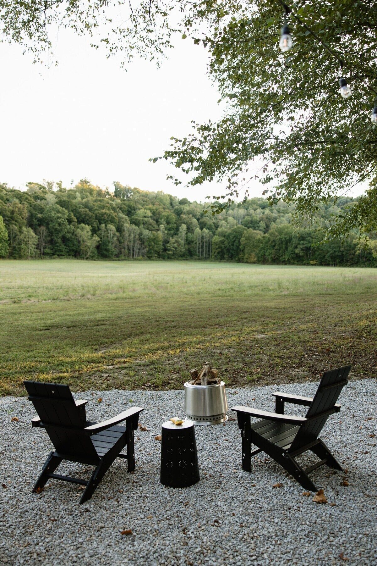 Two Adirondack chairs facing a fire pit on a gravel surface with a forested field in the background.