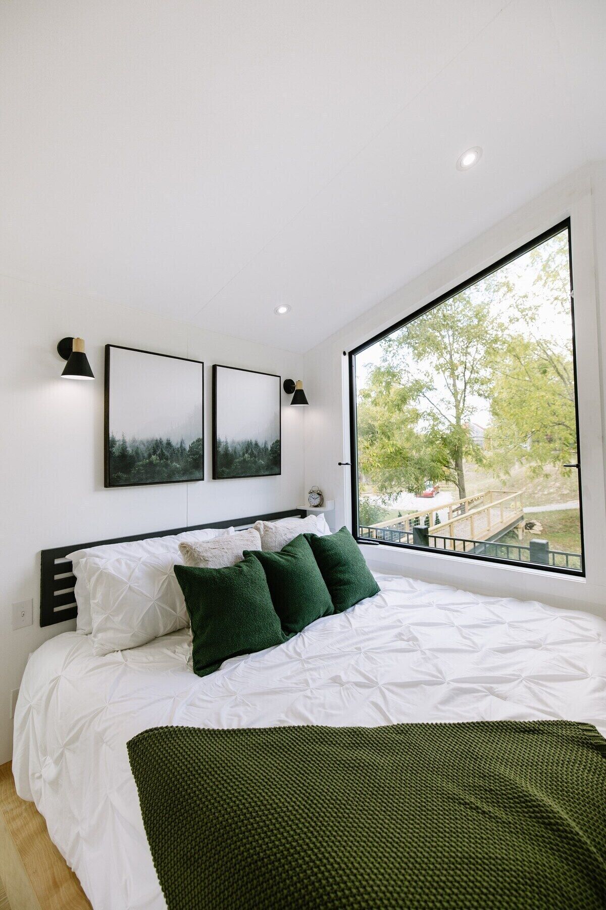 A modern bedroom with a large bed, two wall-mounted picture frames, and a view of a park through a large window.