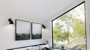 A modern bedroom with a large bed, two wall-mounted picture frames, and a view of a park through a large window.