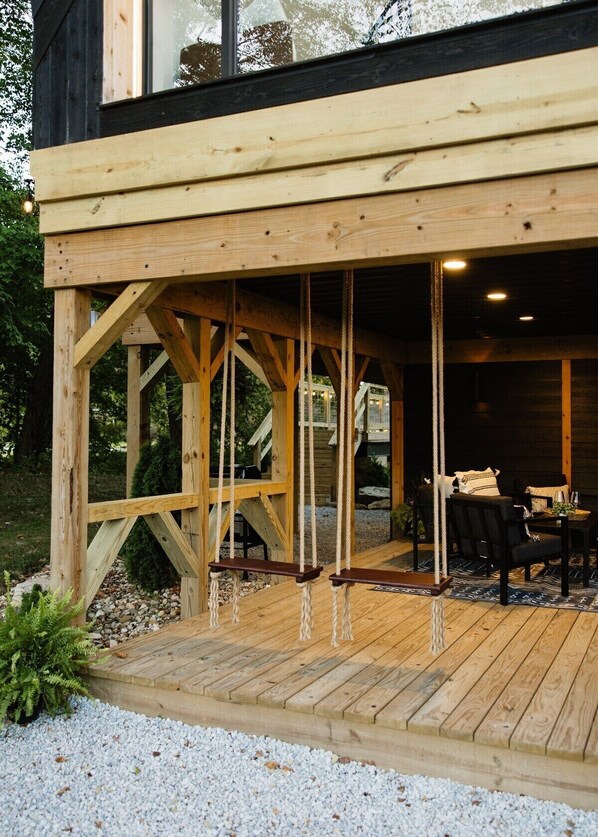 A wooden deck with a swing set, a covered seating area with tables and chairs, and a gravel pathway.