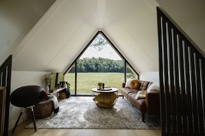 A cozy loft bedroom with a large window overlooking a green field. It features a brown leather sofa, a round coffee table, and a wicker chair.