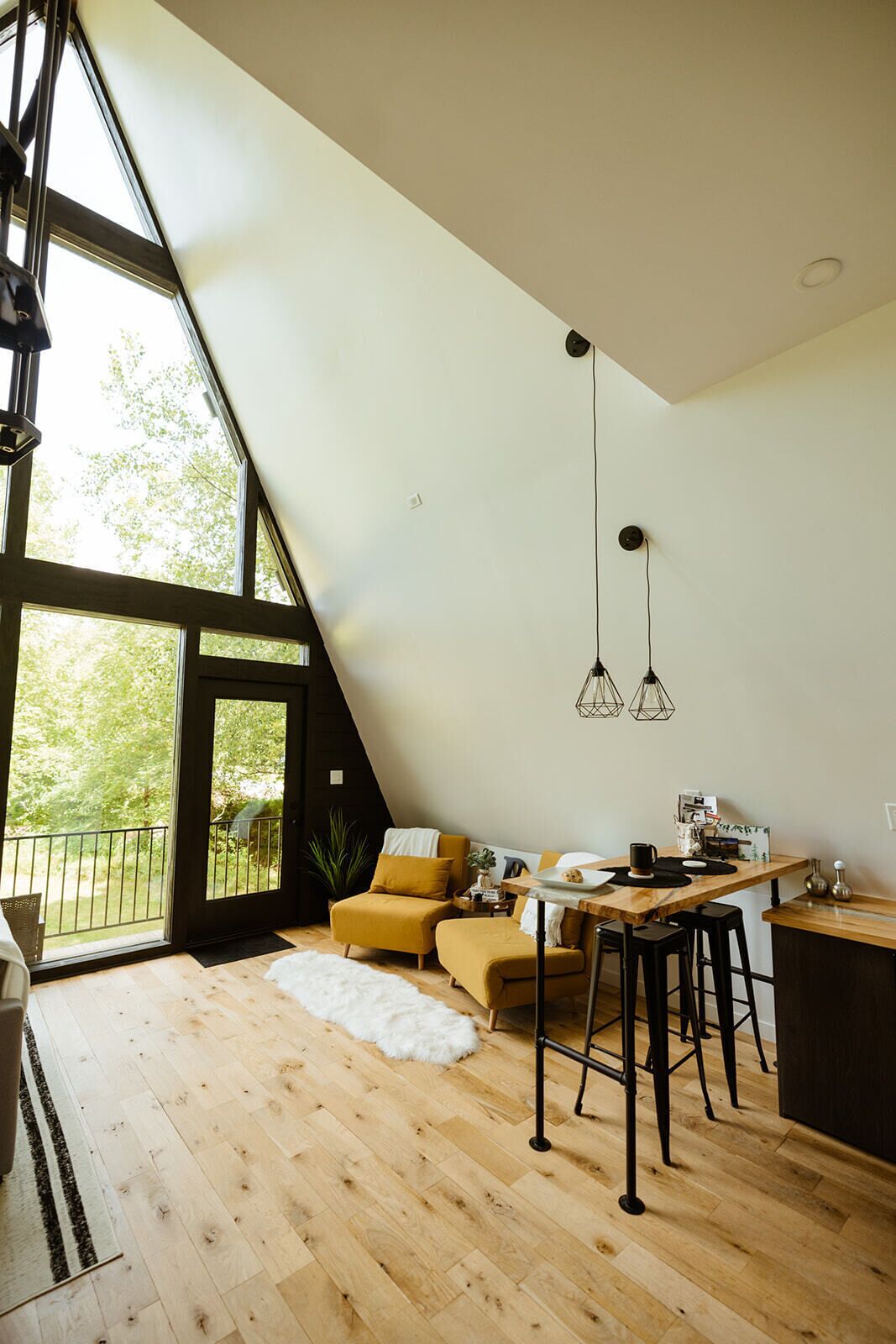 Un loft moderno con techo inclinado, una zona de comedor con mesa y sillas de madera, un sofá con cojines amarillos y un ventanal que ofrece vistas al verdor.