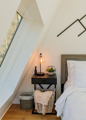 Una camera da letto con soffitto spiovente, un comodino con una lampada, una piccola pianta e un cesto.