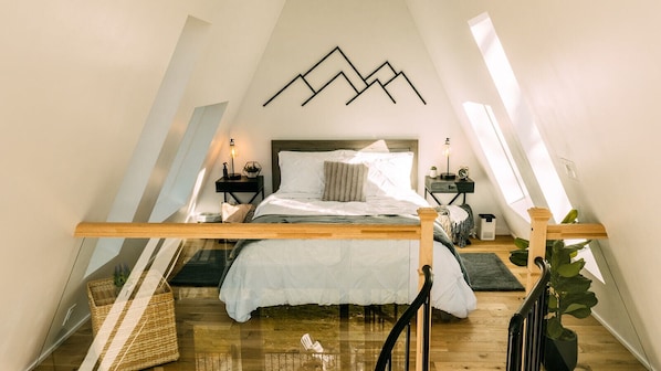 A bedroom with a sloped ceiling, a bed with white bedding, two bedside tables with lamps, and a wall-mounted geometric decoration.