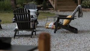 Une terrasse en bois, agrémentée de chaises Adirondack et d’un fauteuil de relaxation, entourée d’arbres et bordée d’un chemin de gravier.