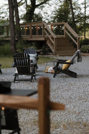 Un terrazzo in legno con poltrone Adirondack e una chaise longue, circondato da alberi e un vialetto sterrato.