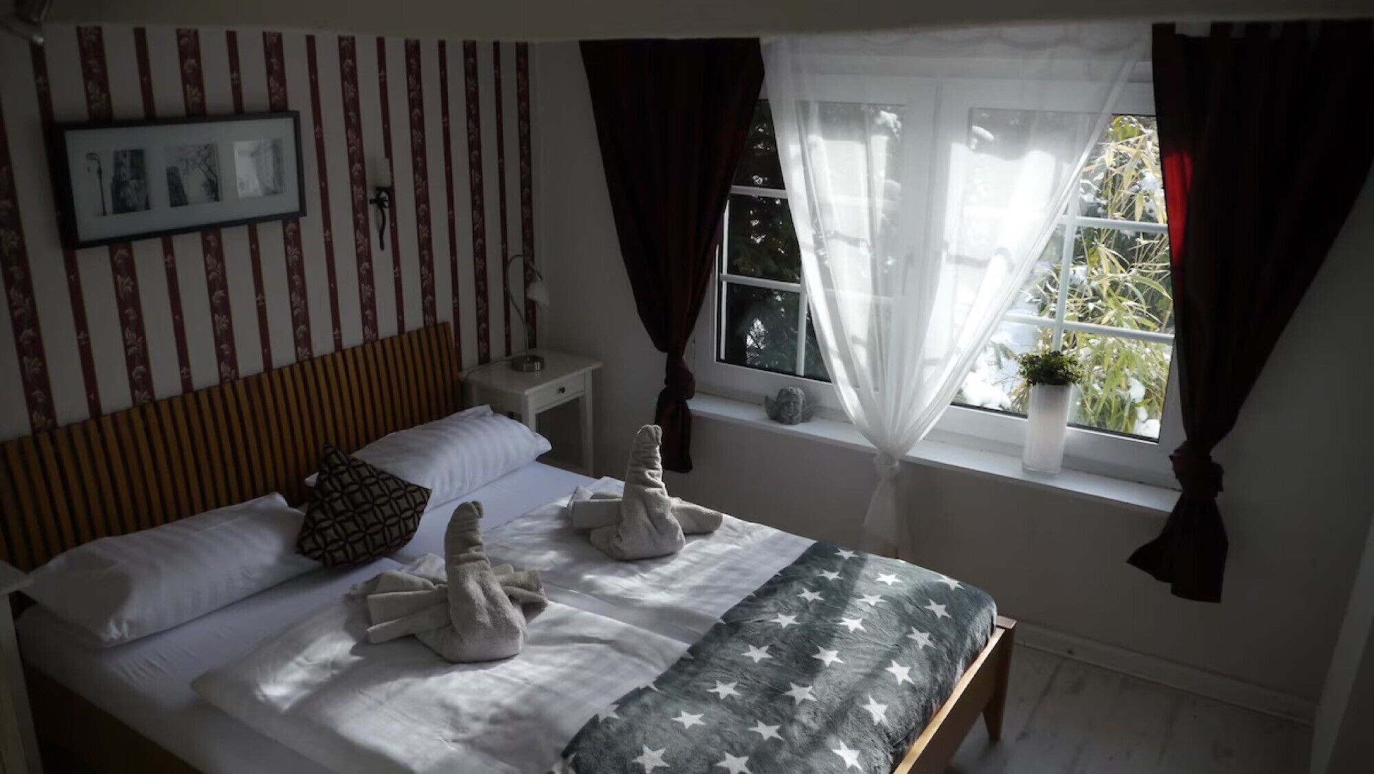 A bedroom with a bed, pillows, a bedside table, a window with curtains, and a wall with vertical stripes.
