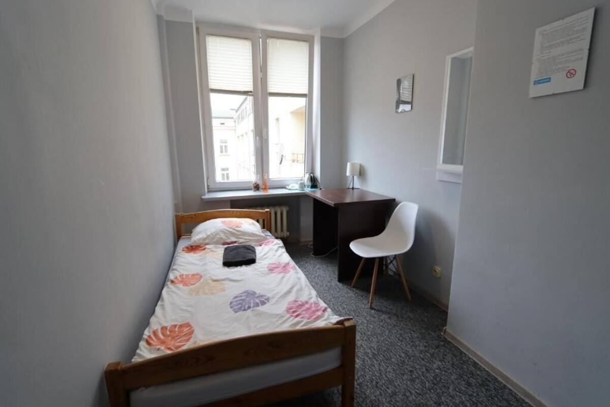 A small, single-bed room with a wooden bed, a desk, and a chair. There is a window with blinds, a radiator, and a notice on the wall.