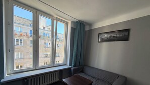 A room with a radiator, a window with curtains, a sofa, a table, and a cityscape painting on the wall. - Fantastic Inn Warsaw - Kopernika (Warsaw)