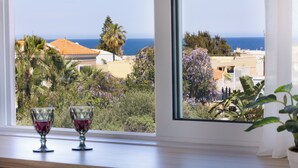 A window with two wine glasses on a wooden ledge, overlooking a coastal view with buildings and greenery. - Sueno Studios and Apartments (Chania)