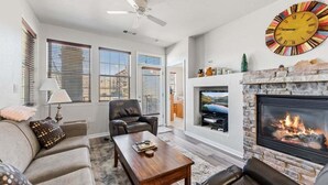 A living room with a fireplace, a ceiling fan, and a large wall clock. - Ten Mile B-102 Ground floor 2 bedroom with private patio upscale modern interior (Granby)