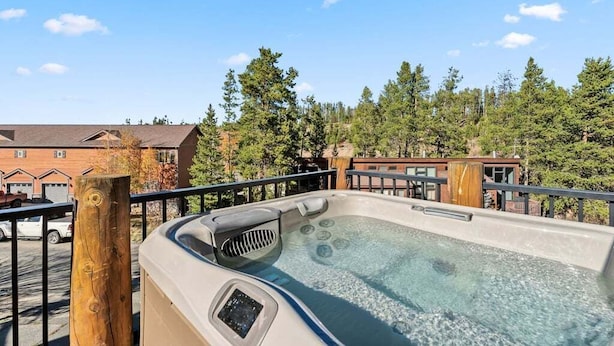 Un jacuzzi en una terraza con baranda de madera, rodeado de árboles y edificios al fondo.