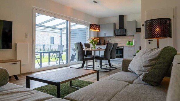 A modern living room with a sofa, a coffee table, and a dining area with chairs and a table. - Ferienwohnung am See in Harkebrügge mit Sauna, WLAN, Terrasse & Weber-Gasgrill (Barßel)