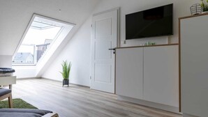 A modern living room with a sloped ceiling, a flat-screen TV mounted on the wall, a wooden floor, and a large window providing natural light. - Ferienwohnung am See in Harkebrügge mit Sauna, WLAN, Terrasse & Weber-Gasgrill (Barßel)