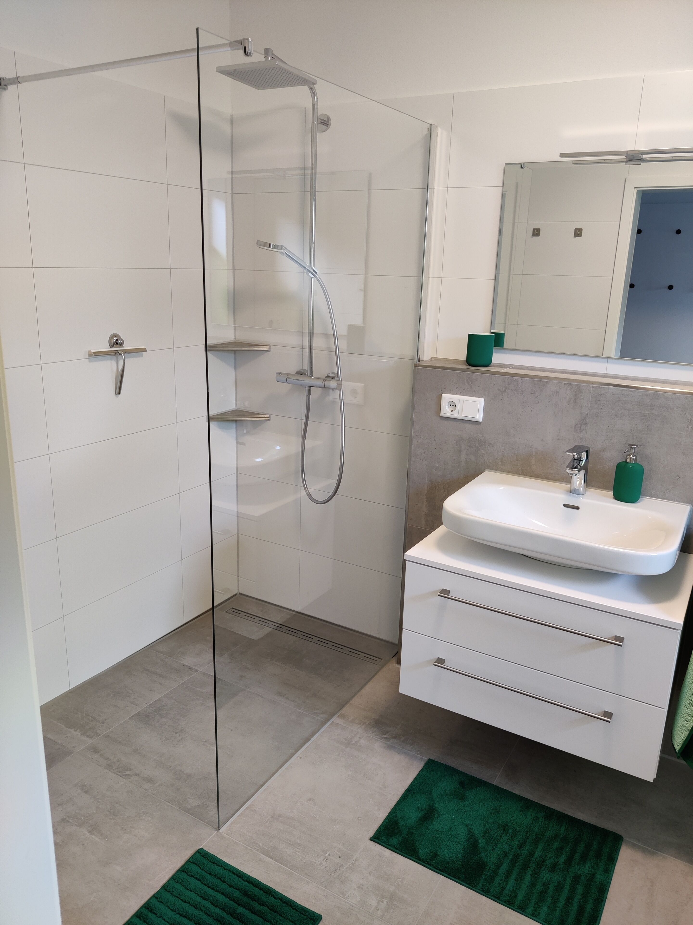 Ein modernes Badezimmer mit einer Duschkabine aus Glas, einem weißen Waschbecken mit grünem Seifenspender und einem Spiegel darüber.