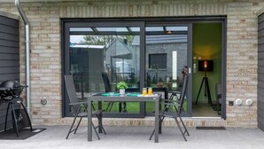 A modern patio with a glass door leading to a room, a table with chairs, a grill, and a tripod with a camera. - Ferienwohnung am See in Harkebrügge mit Sauna, WLAN, Terrasse & Weber-Gasgrill (Barßel)