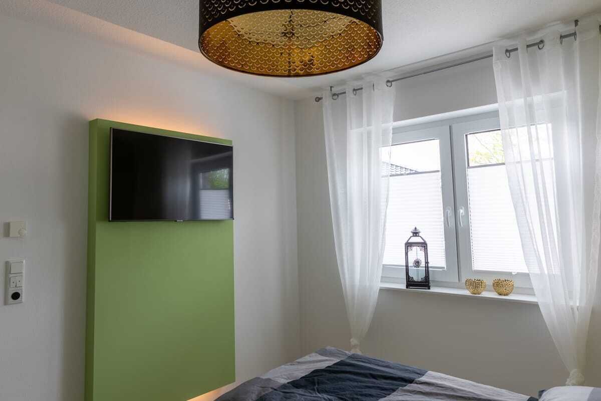 Ein Schlafzimmer mit einem Bett, einem an der Wand montierten Fernseher, einem Fenster mit Vorhängen und einer Laterne auf der Fensterbank.