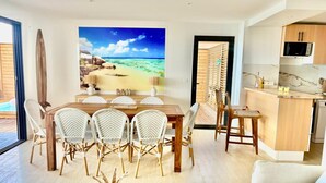 A modern living space with a dining area, kitchen, and a large beach-themed artwork. - Villa Coralia — Le luxe les pieds dans l’eau (Oyster Pond)
