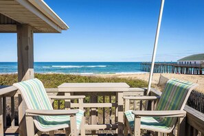 A wooden deck with two striped chairs and a table, overlooking a beach with a pier.
