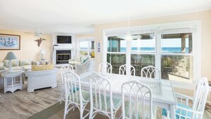 A bright living room with a glass table, white wicker furniture, a fireplace, and a large window overlooking the ocean. - Oceanfront Private Beach Access & Community Pool! (Southern Shores)