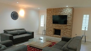 A modern living room with a stone fireplace, a flat-screen TV, and grey sofas. - Pioneer Guest House (DURBAN)