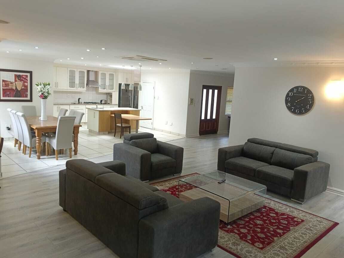A modern living room with grey sofas, a glass coffee table, and a red patterned rug. There is a dining area with a wooden table and chairs, and a kitchen in the background.